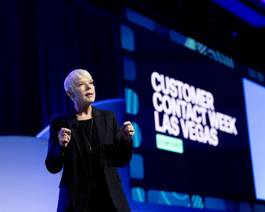 A person with short blonde hair speaks on stage at Customer Contact Week Las Vegas, with a large event sign visible in the background.