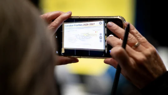 A person takes a photo of a presentation slide titled “Executive Priorities 2026-2027” with a smartphone, while holding a stylus.