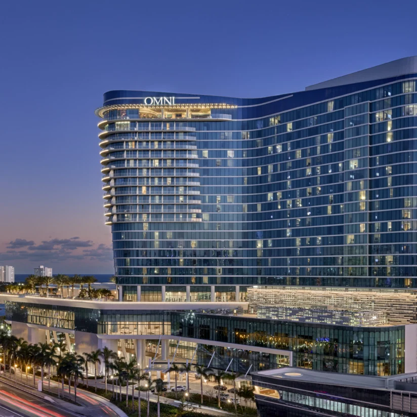 Modern high-rise hotel with curved glass facade, lit at dusk, surrounded by palm trees and busy streets with city buildings in the background.
