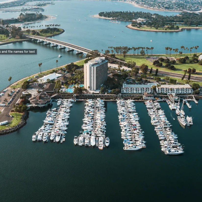 Aerial view of a marina with many docked boats, a tall hotel, a bridge, green spaces, and water surrounding the area.