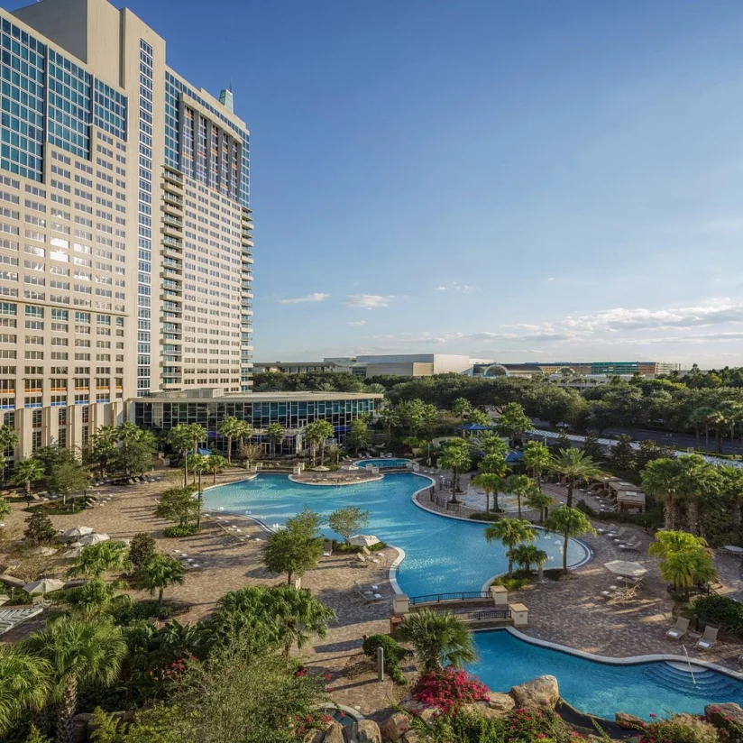 High-rise hotel with many windows overlooks a large, curvy outdoor pool surrounded by palm trees, lounge chairs, and landscaped gardens on a sunny day.