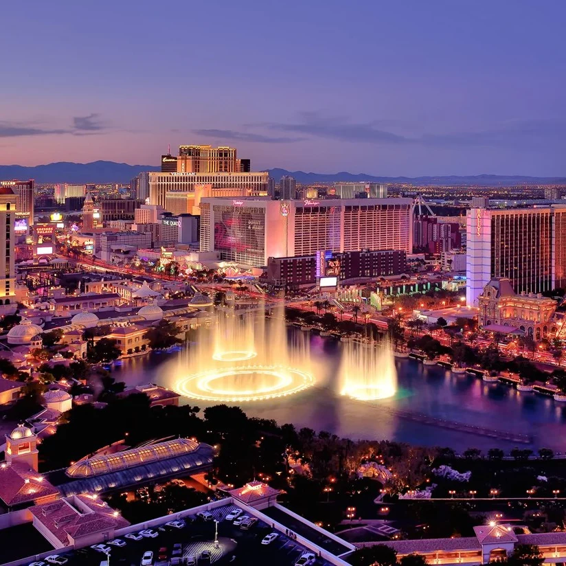 Aerial view of the Las Vegas Strip at dusk, featuring hotels, casinos, and the Bellagio fountain display with city lights illuminating the scene.