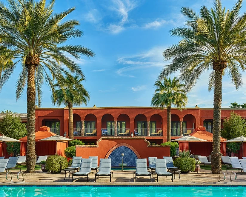 Poolside view with lounge chairs, umbrellas, tall palm trees, and a red stucco building with arches and a blue fountain in the center under a clear blue sky.